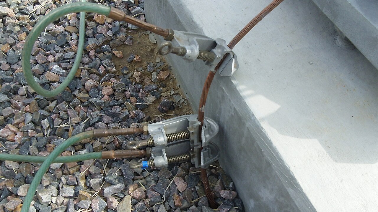 A temporary earthing clamp attached to an optical ground wire cable running down a concrete substation foundation, with the stranded metallic outer layers of the cable clearly visible.
